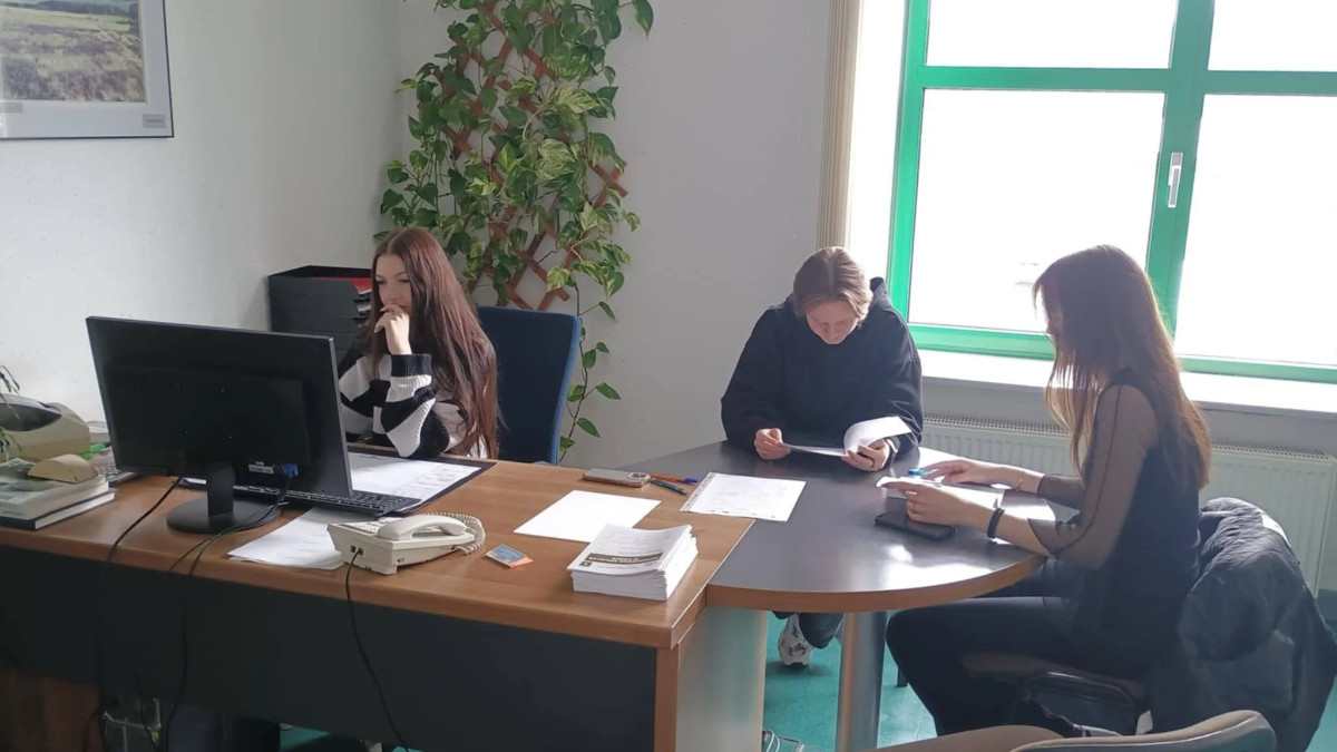 Troje uczniów w trakcie praktyk. Dziewczyna pracuje przy komputerze. Dwoje pozostałych czyta dokumenty
