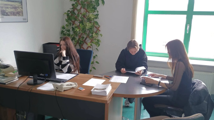 Troje uczniów w trakcie praktyk. Dziewczyna pracuje przy komputerze. Dwoje pozostałych czyta dokumenty