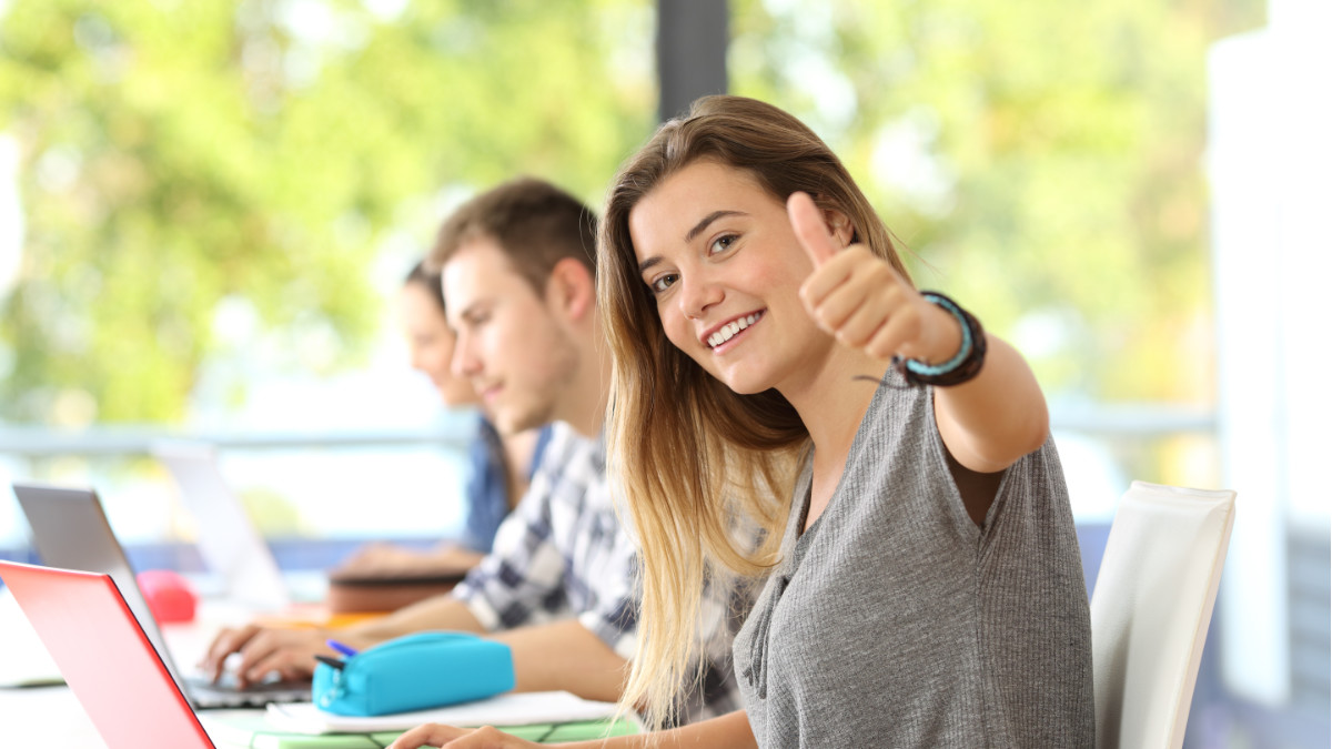 Trzy osoby siedzą przy stole na którym leżą laptopy. Dziewczyna na pierwszym planie przy ma podniesioną rękę i kciuk skierowany do góry