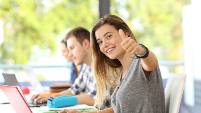 Trzy osoby siedzą przy stole na którym leżą laptopy. Dziewczyna na pierwszym planie przy ma podniesioną rękę i kciuk skierowany do góry